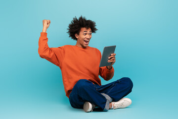 A young black man with curly hair is seated on the floor, excitedly celebrating while holding a...
