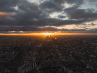The image shows an aerial view of London at sunset, highlighting silhouetted skyscrapers in Canary Wharf against a vibrant sky with dramatic clouds.