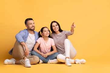 A loving family of three shares a joyful moment against a bright yellow background. The parents sit closely with their daughter, who points at copy space while they all smile