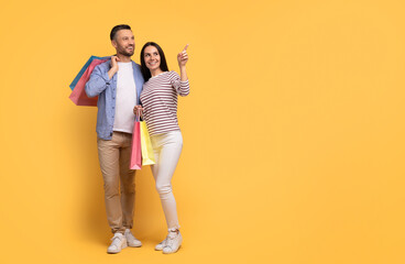 A joyful couple stands together, holding colorful shopping bags. They share a moment of affection, with the woman pointing playfully as they enjoy their outing, copy space