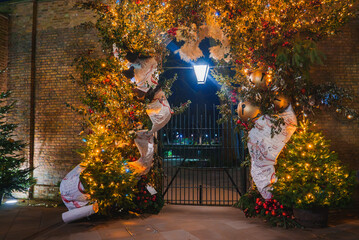 A decorated archway with golden lights, greenery, and baubles against a brick wall. A wrought iron...