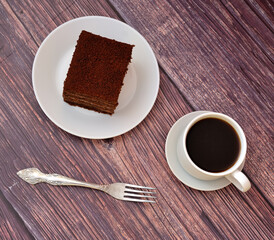 Layered sponge chocolate cake on white plate with cup of hot black coffee on wooden table.