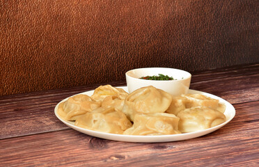 A large plate of freshly cooked meat manti and a cup of tomato spicy sauce on a wooden table.