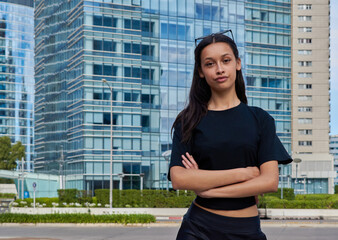A woman with sunglasses on her head, dressed in a black T-shirt and dark pants, stands with her arms crossed in front of a modern office building with glass windows.