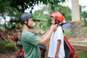A asian father adjusts a child helmet for safety before riding a red electric scooter together outdoors. © Iqy