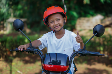 A young asian boy in a red helmet gives a thumbs up while sitting on a electric bike in outdoor