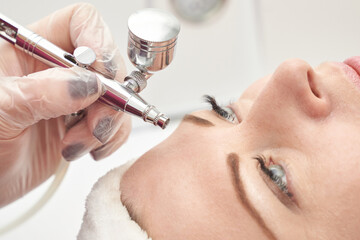 Caucasian female receiving airbrush facial treatment in beauty salon with gloved hands applying...