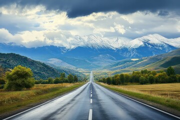 Fototapeta premium Scenic highway through autumnal valley towards snow-capped mountains under a dramatic sky.