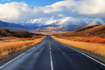 Scenic highway road through autumnal landscape towards snow-capped mountains under a partly cloudy sky.