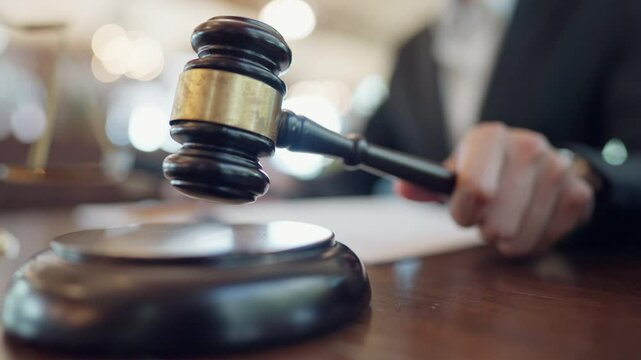 A person is holding a gavel and standing in front of a judge's bench. The person appears to be a judge or a lawyer, and the scene suggests a formal setting