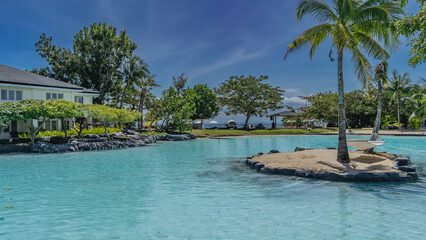 A recreation area in a tropical resort. There is a tiny islet in the middle of the aquamarine swimming pool. The hammock is suspended between the trunks of palms.Ocean, sun umbrellas in the distance. 