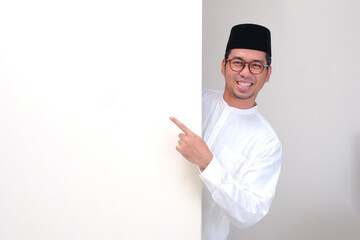 A muslim man peeking out behind wall smiling and pointing at it