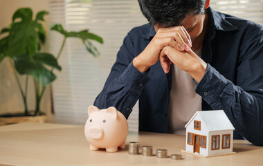 Stressed man holding his forehead, surrounded by a piggy bank, coin stacks, and a house model....