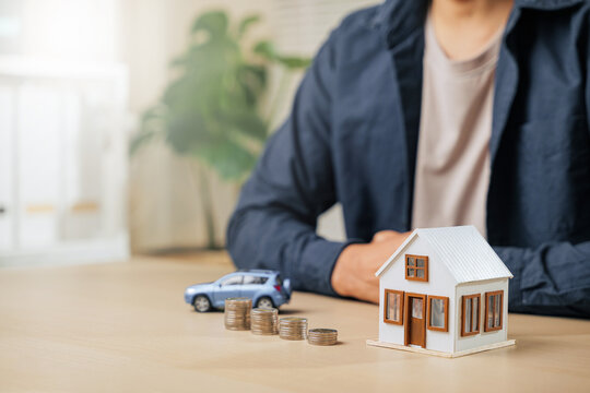 A person sits with hands clasped near a small model car, house, and coin stacks. Highlights finance, saving, property investments, auto and home loans, installment payments