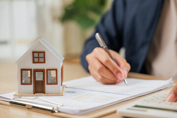 A man writes a signature on house insurance documents, using a calculator for tax and property details. Counting payments and rent by model house, he finalizes the price