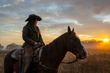 Woman Wearing Cowboy Hat Riding Horse and Looking Off At Golden Hour of Sunset With Cloudy Sky