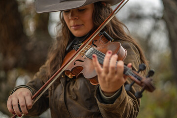 Close Up of Woman Wearing Cowboy Hat and Playing Fiddle Violin In Front of Blurred Trees © Lone Star Stock