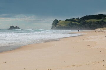 Cloudy day at the beach. Gentle waves crash on the shore as a person walks along the sand. Coastal scenery. OPOUTERE, COROMANDEL PENINSULA, NEW ZEALAND
