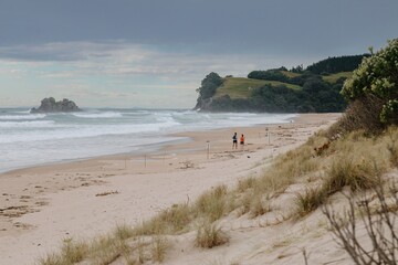 Cloudy day at the beach. Couple walks along the shore, amidst powerful waves. Coastal scenery. OPOUTERE, COROMANDEL PENINSULA, NEW ZEALAND