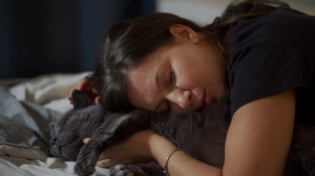 Adult 44 year old woman and gray domestic cat lying on bed in bedroom. Happy woman hugging and kissing her cat, stroking his belly.