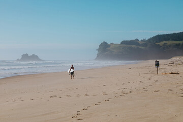Surfer walks along a tranquil beach, carrying a surfboard. Coastal scene with calm waves. OPOUTERE, COROMANDEL PENINSULA, NEW ZEALAND