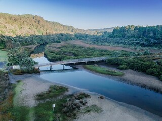 Wooden boardwalk bridge over tranquil waterway, reflecting in still water. Nature preserve, early morning light. OPOUTERE, COROMANDEL PENINSULA, NEW ZEALAND