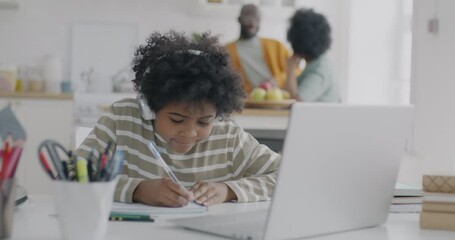 African American schoolboy learning in virtual classroom listening to teacher with headphones and laptop while parents talking in kitchen. Education and people concept. - Powered by Adobe