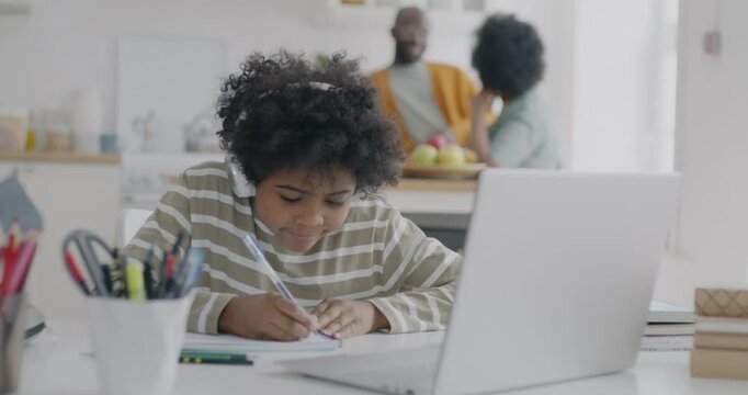 African American schoolboy learning in virtual classroom listening to teacher with headphones and laptop while parents talking in kitchen. Education and people concept. - Powered by Adobe