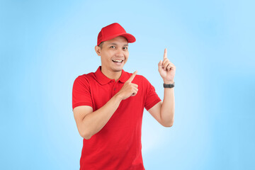 An enthusiastic Asian courier in a red uniform and cap points upwards with a bright smile, highlighting a service offer or promotional message in a studio setting with a blue background.