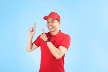 An enthusiastic Asian courier in a red uniform and cap points upwards with a bright smile, highlighting a service offer or promotional message in a studio setting with a blue background.