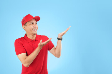 An enthusiastic Asian courier in a red uniform and cap points upwards with a bright smile, highlighting a service offer or promotional message in a studio setting with a blue background.