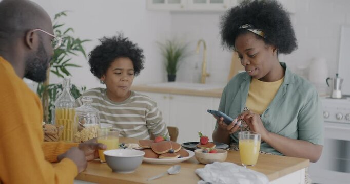 Happy African American woman having breakfast with husband and son and taking pictures of food with smartphone camera in kitchen at home. Family lifestyle and modern technology concept.