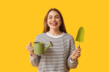 Beautiful young woman with watering can and gardening shovel on yellow background