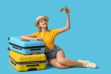 Beautiful young woman with toy airplane and suitcases sitting on blue background. Travel concept