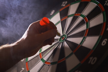 Hand Aiming Red Dart at Bullseye on Dartboard in Smoky Background