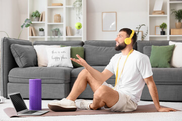 Sporty young man with headphones, apple and laptop sitting on yoga mat at home