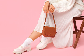 Young African-American woman with stylish brown handbag sitting on chair against pink background