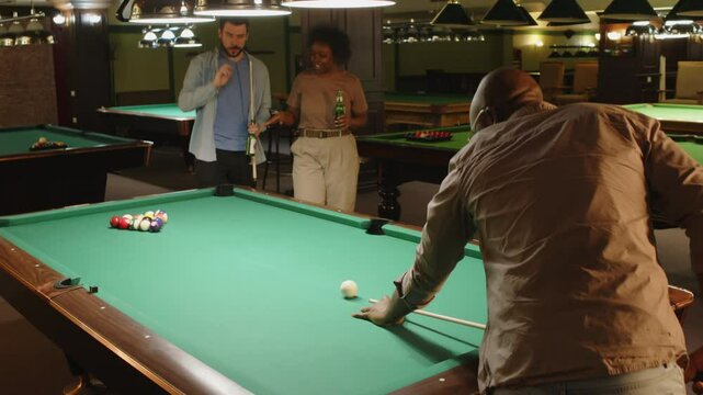 Back view of African American man aiming at cue ball while starting playing pool game with diverse friends in bar
