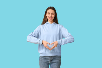 Beautiful young woman making heart gesture on blue background
