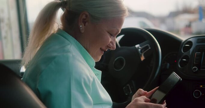 A smiling nurse on a break in her car types a message on her cell phone