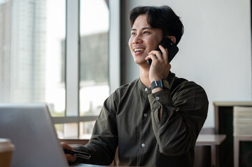 A positive Asian businessman talking on the phone while working remotely from a coffee shop.