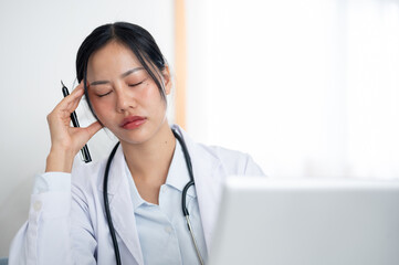 Overworked, tired Asian female doctor working at her desk in the office, suffering from a headache