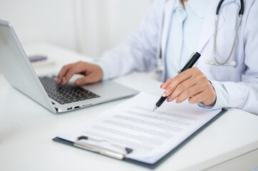 A close-up of a doctor working at her desk, typing on the laptop keyboard, reading medical paper.