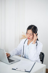 Overworked, tired Asian female doctor working at her desk in the office, suffering from a headache