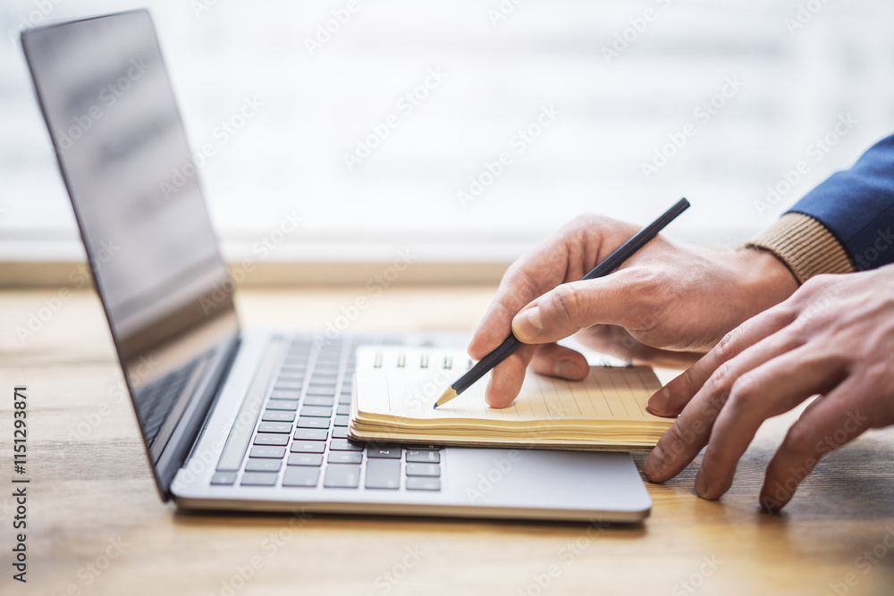 Poster focused view of a man's hand as he writes in a notepad on a modern laptop, background gently blurred