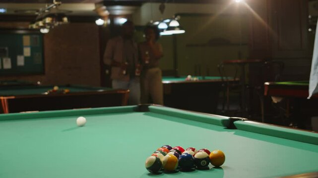 Full medium shot of excited Caucasian man preparing baize table for pool game and breaking billiard balls while his African American friends reaching him with bottles of beer in pub