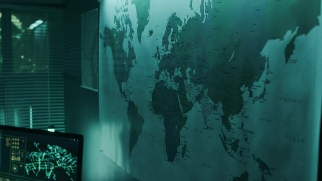Medium tilting up shot of computer with surveillance program on screen, and world map on wall above desk of intelligence officer at military operations center, in dim blue light at night