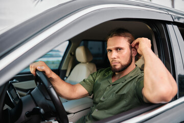 Man sitting in a car, looking thoughtful while adjusting his hair, dressed in a casual green shirt against a blurred urban background