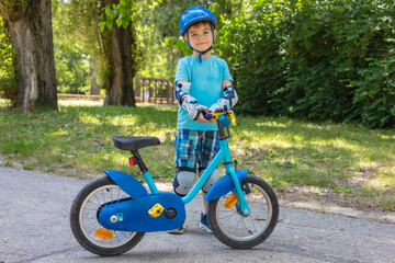 Little boy holding his bicycle. Summer, child likes to ride a bike in the park. Healthy lifestyle concept. Children's set of cycling protection - helmet, knee pads, elbow pads, gloves. Safety first.