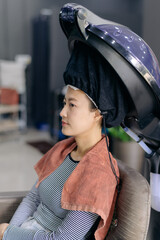 Asian woman sitting in salon chair under hair steamer, wearing striped shirt and towel, in modern...